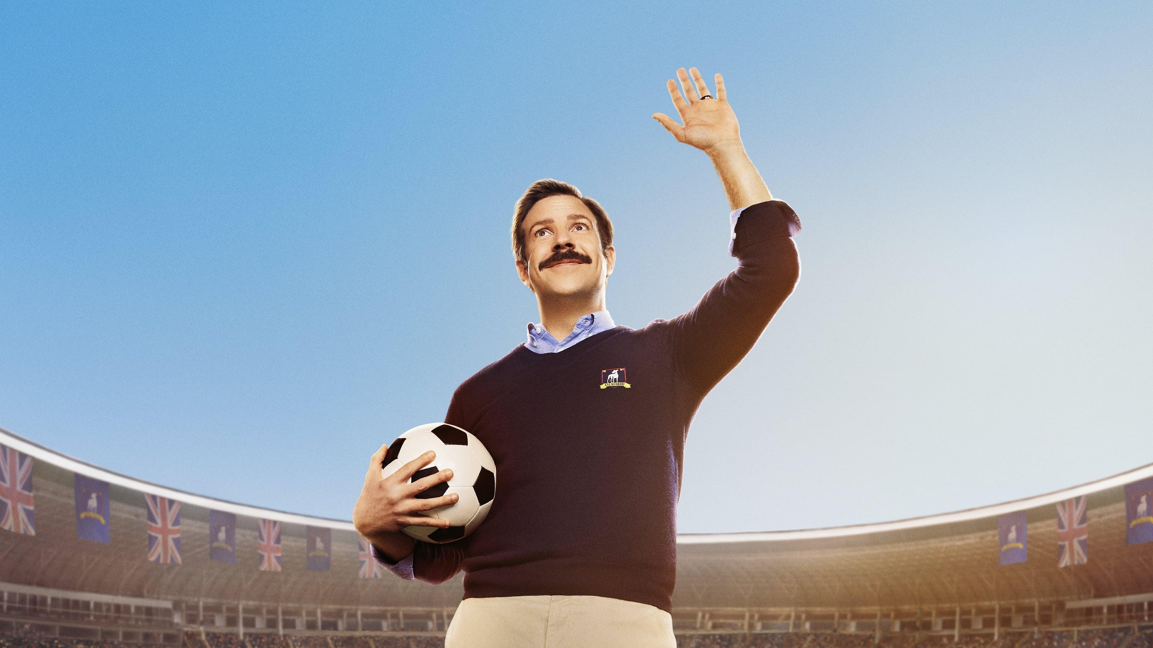 Jason Sudeikis as his character Ted Lasso holding a soccer ball with a raised hand, set against a sunny sky and soccer stadium.